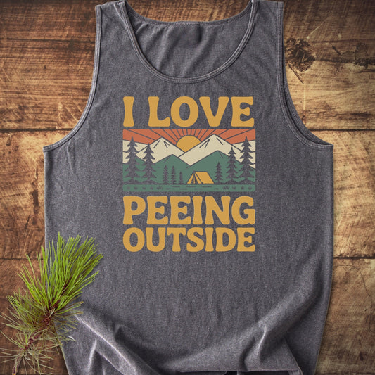 I Love Peeing Outside Tank Top featuring a colorful retro camping graphic with mountains and a tent, displayed on a wooden surface with a pine branch, highlighting sustainable, comfortable US cotton.