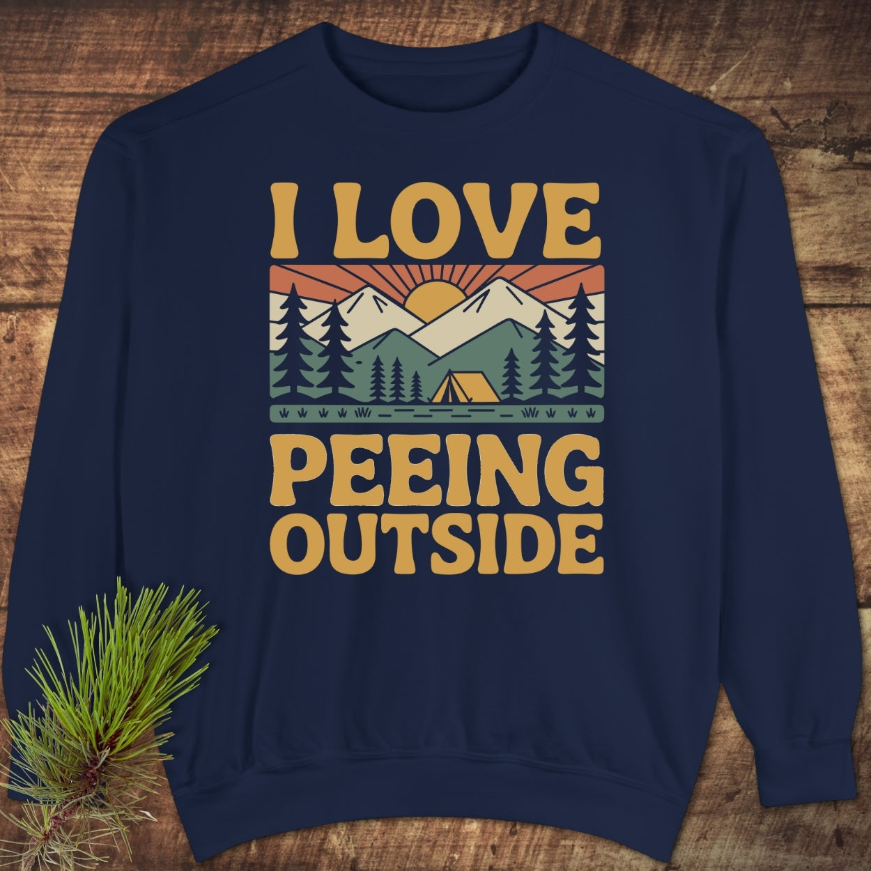 I Love Peeing Outside Sweatshirt featuring a retro sunrise graphic over mountains and trees, laid flat with pine sprigs. Unisex, garment-dyed with relaxed fit, showcasing bold, outdoor-themed text.