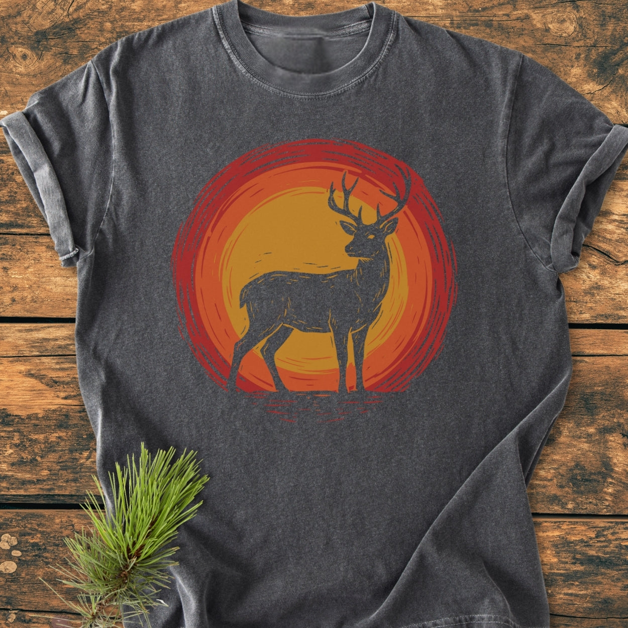 Trail Guardian - Vintage Wash Tee: A dark charcoal-gray T-shirt featuring a stag silhouette against an orange-red sun motif, laid on wood with a pine sprig.