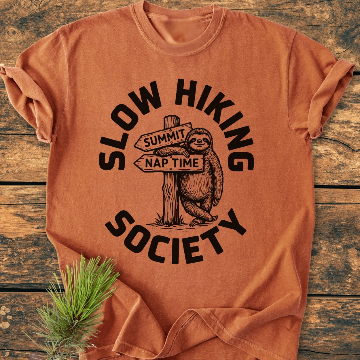 Slow Hiking Society - Vintage Wash Tee featuring a cartoon sloth leaning on a trail sign. Displayed on a wooden surface, it embodies a relaxed fit with durable double-needle stitching.
