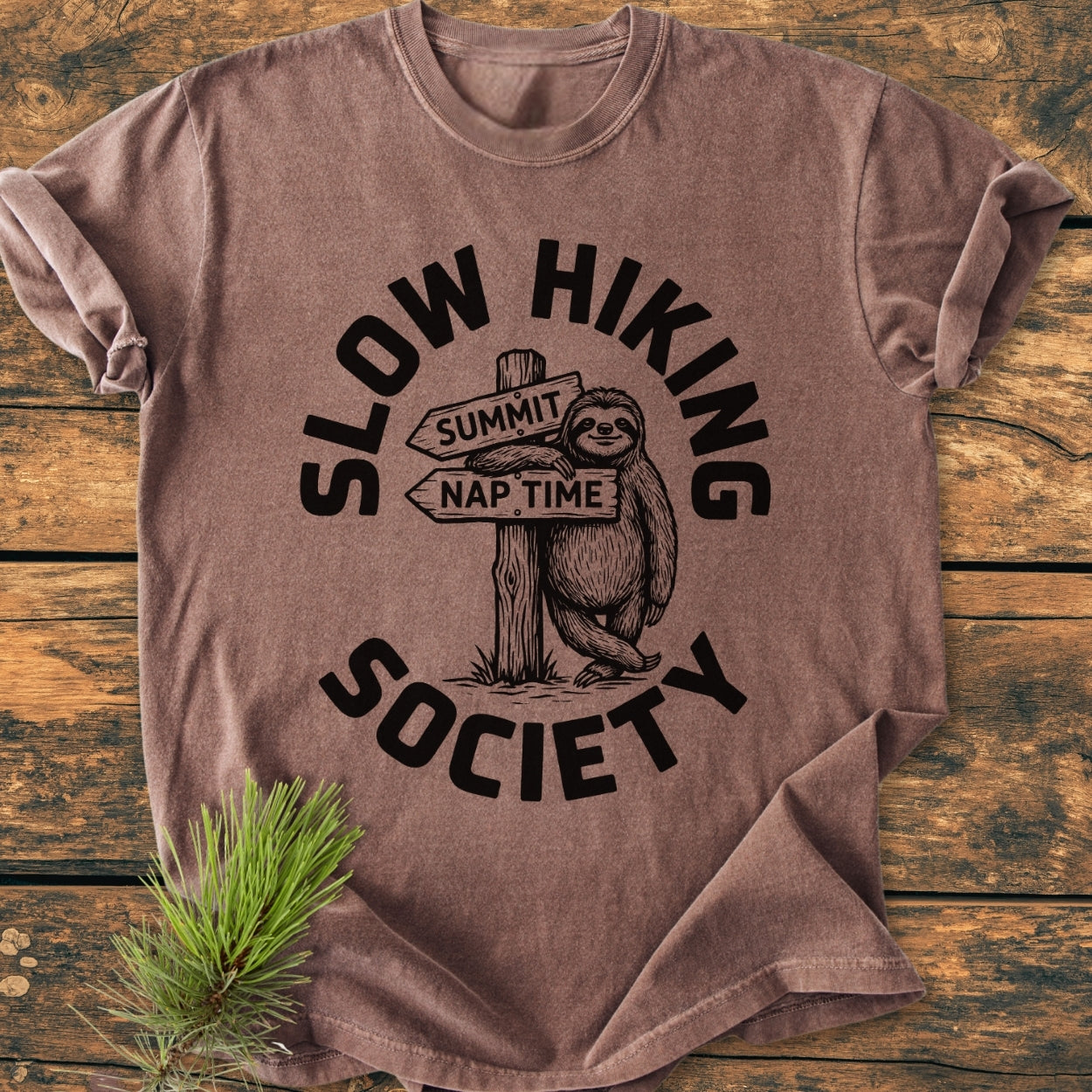 Slow Hiking Society - Vintage Wash Tee featuring a cartoon sloth with a trail-sign. Laid on wood, pine sprig beside. Made of soft, durable, garment-dyed cotton for a relaxed fit.