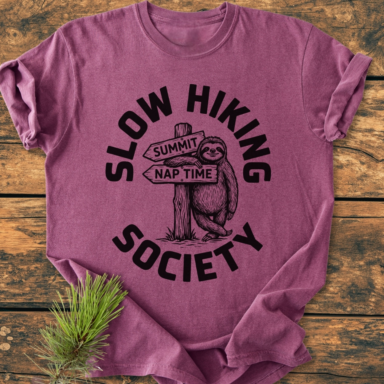 Slow Hiking Society - Vintage Wash Tee features a sloth graphic with signposts 'SUMMIT' and 'NAP TIME,' laid flat on rustic wood with a pine sprig, highlighting its relaxed, cozy design.