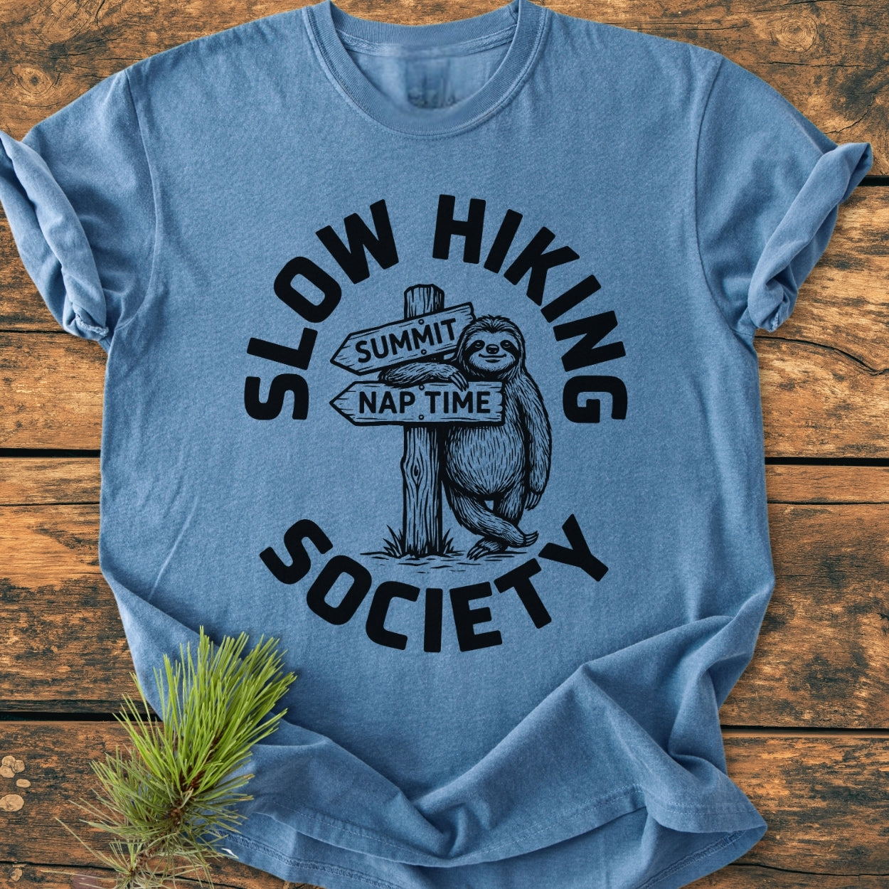Slow Hiking Society - Vintage Wash Tee features a smiling sloth print on a heather-blue T-shirt, laid on rustic wood, highlighting its cozy, relaxed fit and durable construction.