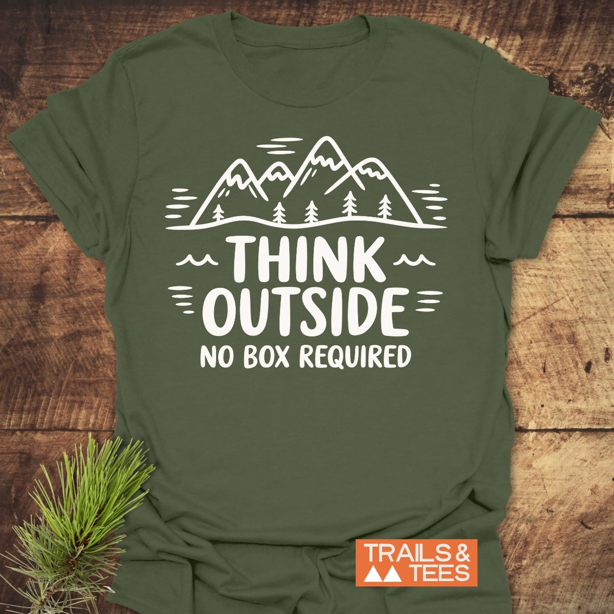Think Outside T-Shirt featuring a white mountain and pine tree design with slogans, laid flat with a pine sprig on a wooden surface, showcasing the Trails & Tees logo.