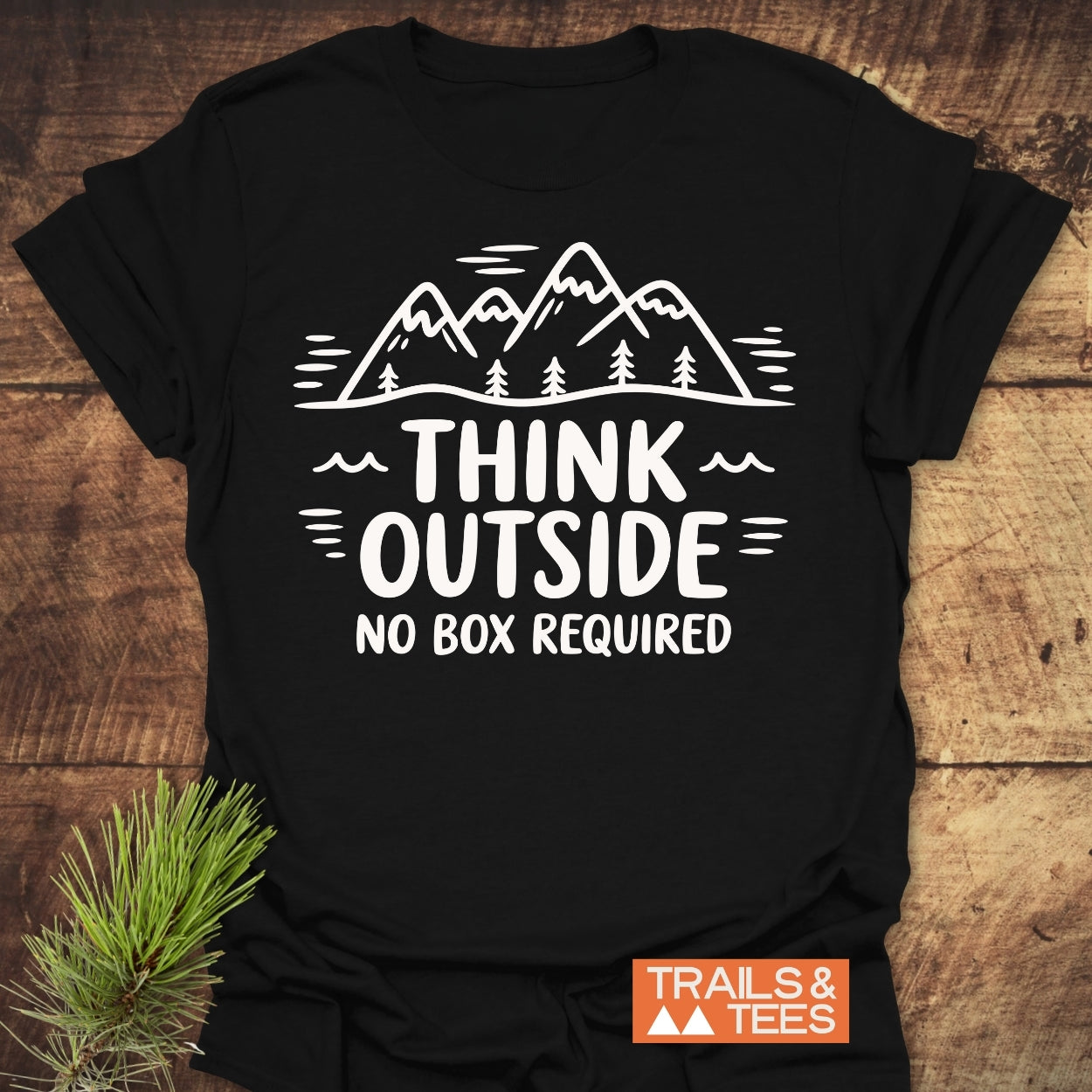 Think Outside T-Shirt on wood surface, featuring a mountain-and-trees graphic with THINK OUTSIDE NO BOX REQUIRED slogan. Includes orange TRAILS & TEES brand tag. Pine sprig accents the outdoor theme.