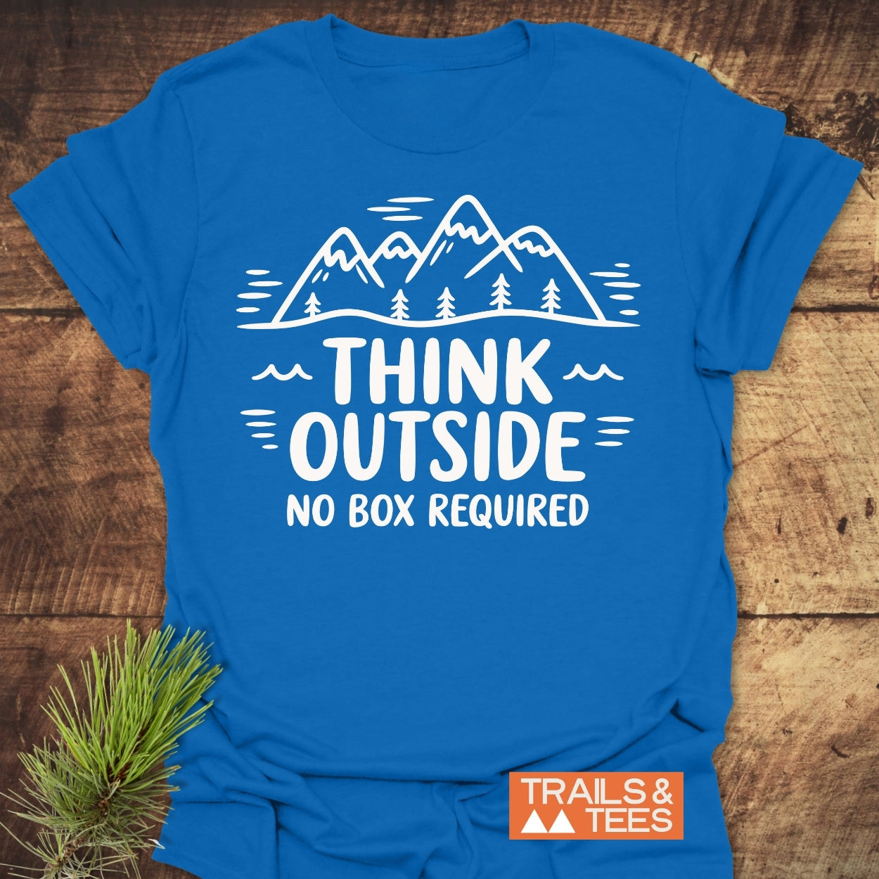 Think Outside T-Shirt featuring a white graphic of mountains and trees with the slogan THINK OUTSIDE NO BOX REQUIRED, laid flat with a pine branch accent and TRAILS & TEES. logo.