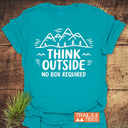The Think Outside T-Shirt features a mountain scene with pines and the slogan THINK OUTSIDE NO BOX REQUIRED in white. A small TRAILS & TEES logo and an evergreen sprig accompany it.