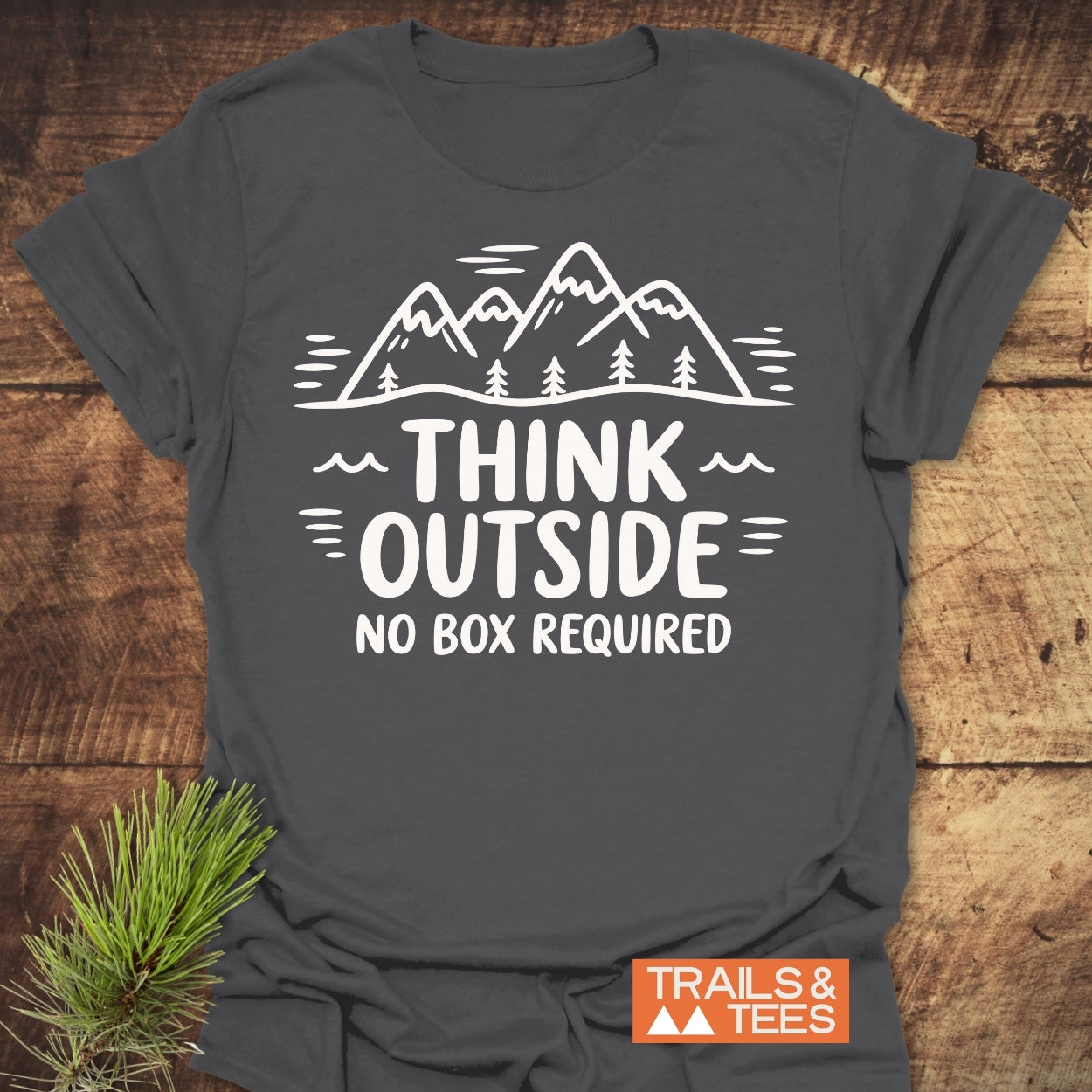 Think Outside T-Shirt featuring a white mountain and pine tree design with “THINK OUTSIDE NO BOX REQUIRED” text. Includes an orange TRAILS & TEES logo. Laid on wood with pine sprig accent.