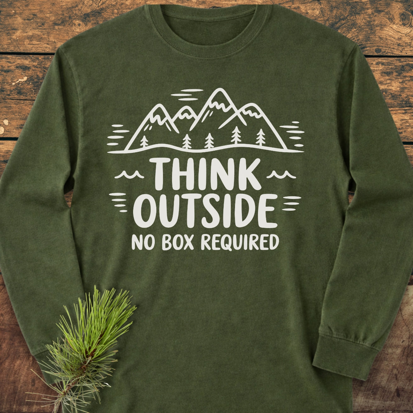 The Think Outside Long-Sleeve Tee features an olive-green design with a mountain-and-pine-tree graphic and the slogan “THINK OUTSIDE – NO BOX REQUIRED,” laid on a wooden surface with a small pine branch accent.