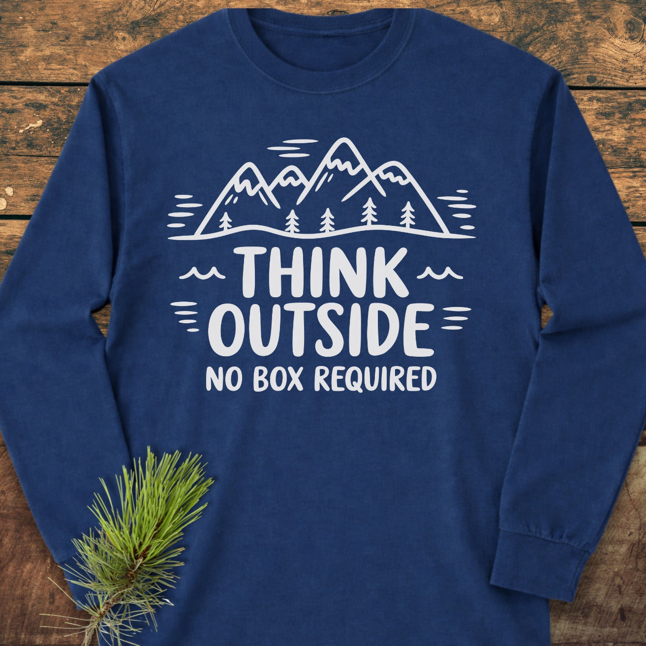 Think Outside Long-Sleeve Tee with white mountain and tree line-drawing and slogan on chest, laid flat on wood with pine sprig nearby. Made of 100% ring-spun cotton for comfort.