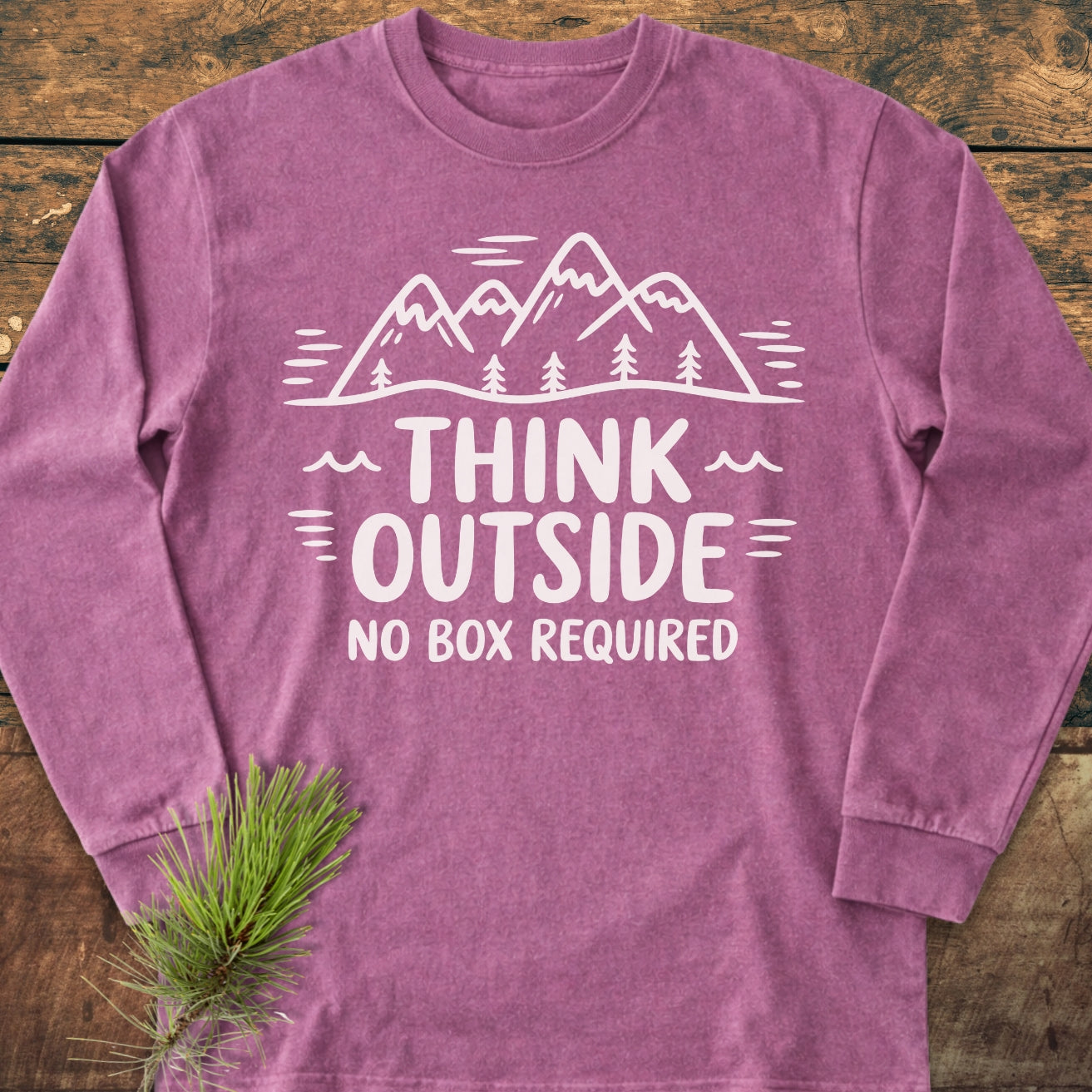 Think Outside Long-Sleeve Tee featuring a mountain-and-tree graphic with the slogan THINK OUTSIDE, NO BOX REQUIRED, displayed on a wooden surface. Enhanced by a pine sprig accent.