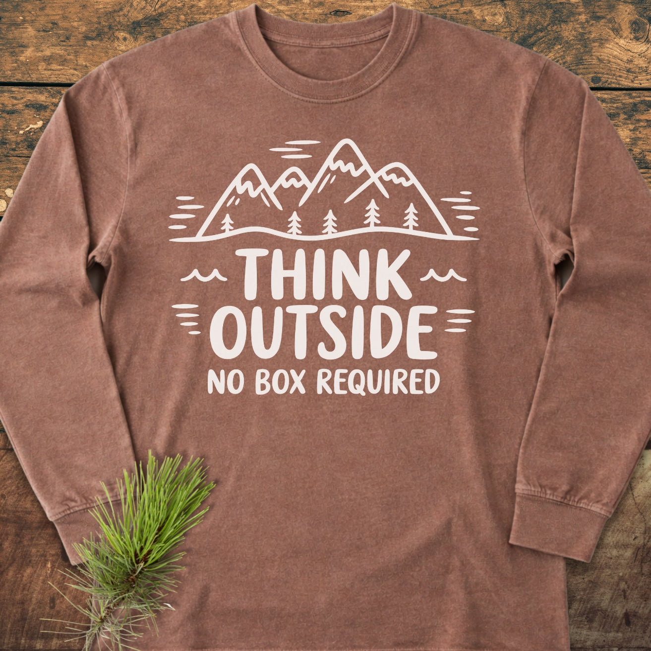 Think Outside Long-Sleeve Tee displayed on wood, featuring a mountain-and-tree graphic with the slogan THINK OUTSIDE NO BOX REQUIRED. A pine sprig accents the soft, garment-dyed, cotton tee.