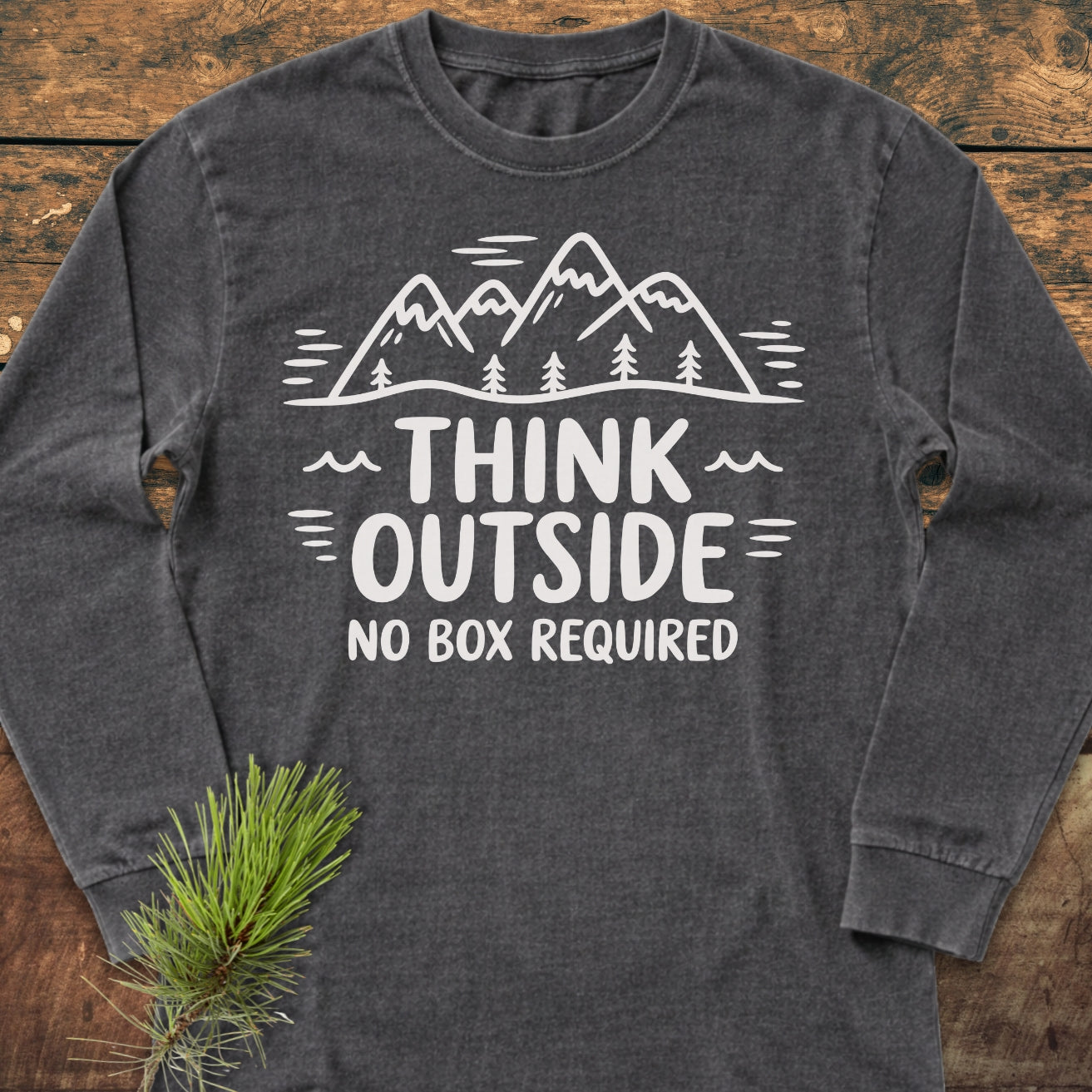 Think Outside Long-Sleeve Tee, featuring a mountain graphic and slogan, laid flat with pine sprig accent. Made from soft, 100% ring-spun cotton for comfort and style.