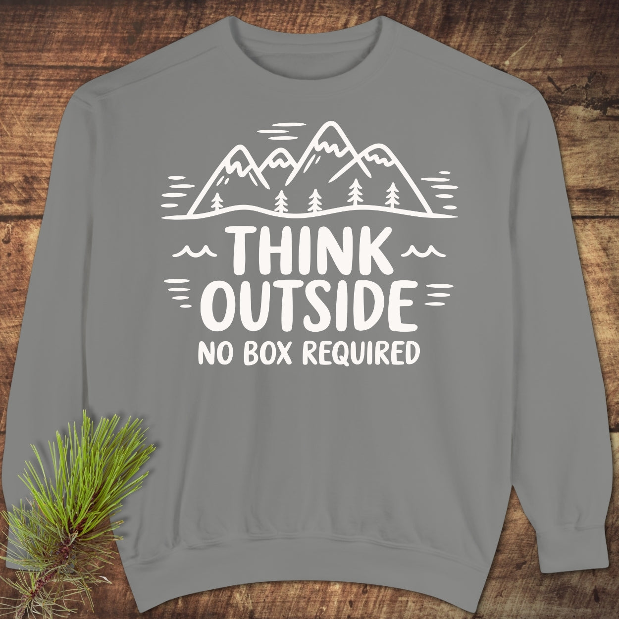 Think Outside Sweatshirt featuring a white mountain-and-tree line drawing and slogan. Displayed on a rustic wood background with a pine sprig, highlighting its relaxed fit and luxurious fabric texture.