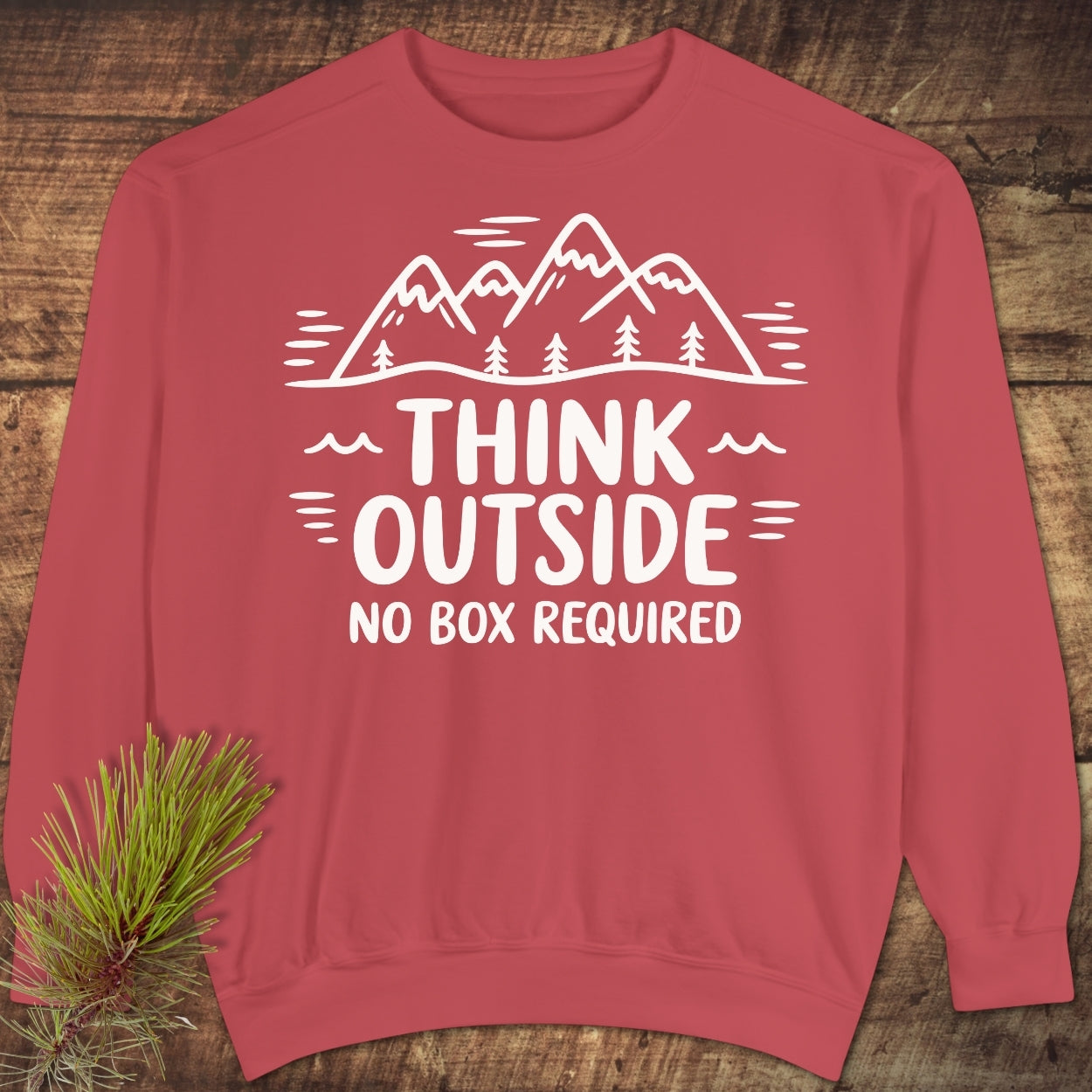Think Outside Sweatshirt with mountain-and-tree graphic and motivational text, displayed on a wooden surface with pine sprig. Features relaxed fit, ring-spun cotton blend, and rolled-forward shoulder design.