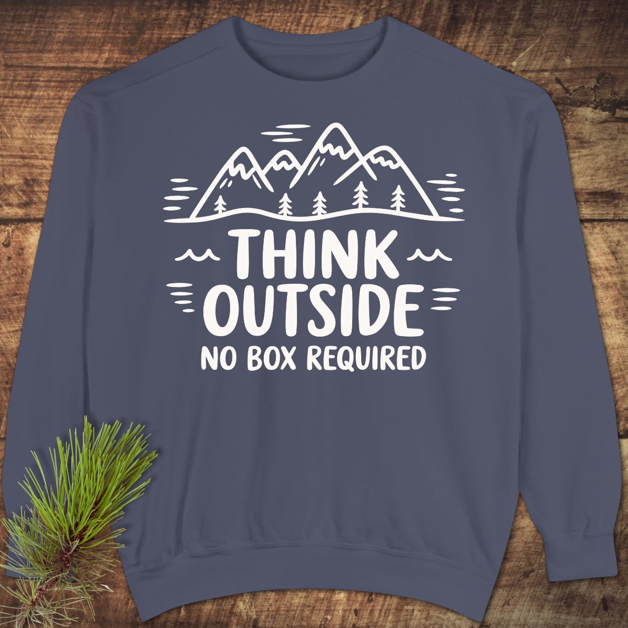 Think Outside Sweatshirt: Slate-blue sweatshirt with a white mountain-and-pine silhouette and slogan. Features include a relaxed fit and rolled-forward shoulder, displayed with a rustic pine sprig accent.