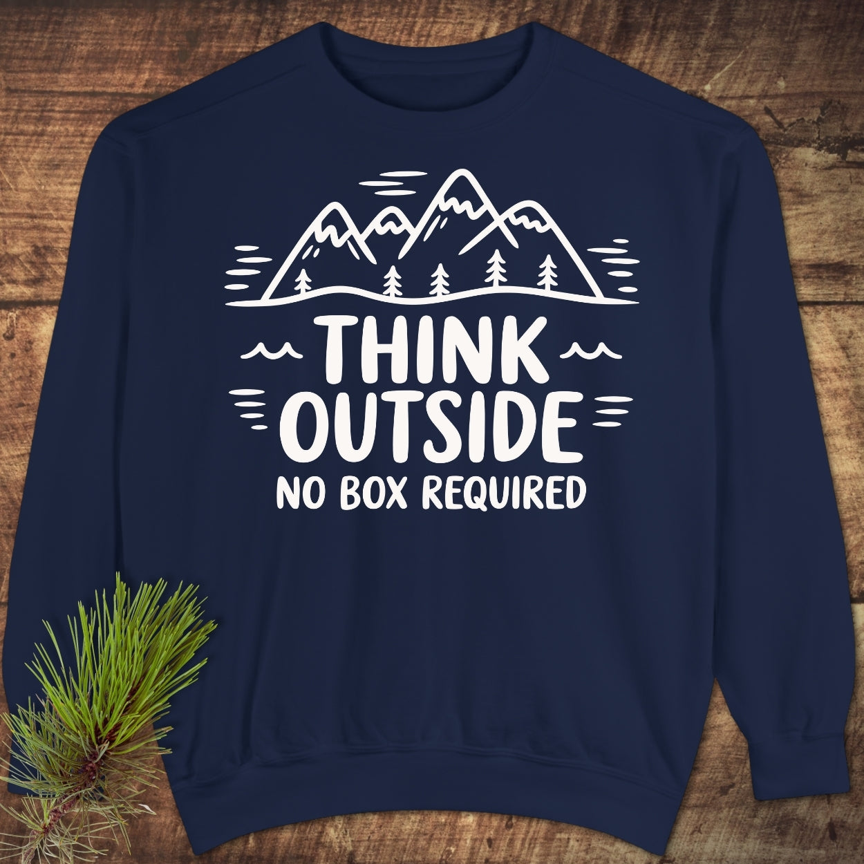 Think Outside Sweatshirt on wooden surface, featuring a white mountain and pine design with THINK OUTSIDE NO BOX REQUIRED slogan; showcases relaxed fit and luxurious fabric blend for comfort.