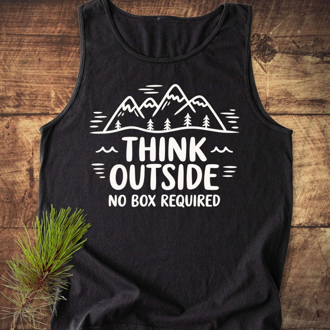 Think Outside Tank Top displayed on wood, featuring a white mountain-and-trees graphic with the slogan “THINK OUTSIDE NO BOX REQUIRED.” It's a black, sleeveless, unisex tank made from sustainable US cotton.