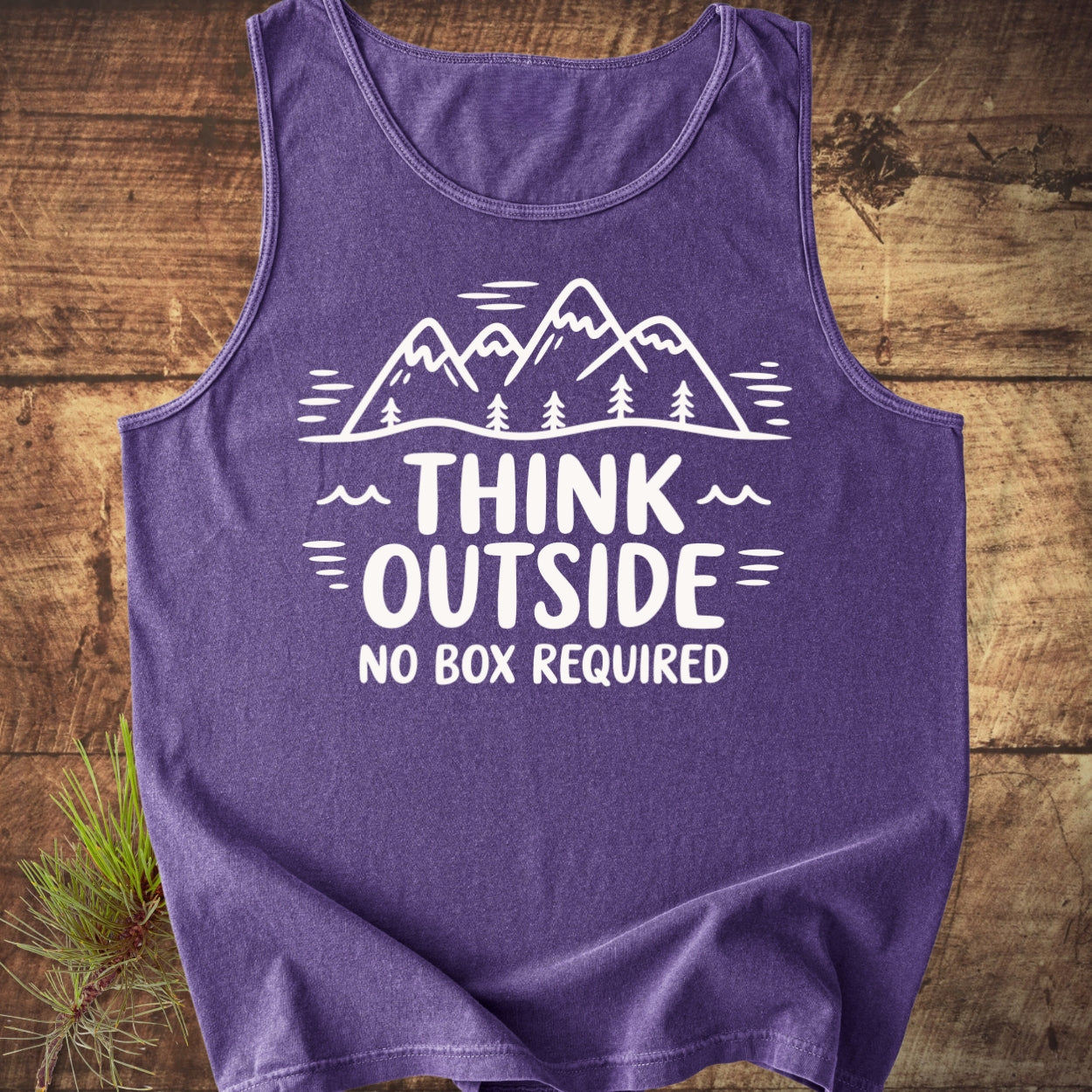 Purple Think Outside Tank Top featuring a white graphic of stylized mountains and pine trees with the slogan, laid flat on wood surface; sprig of pine needles tucked under the edge.