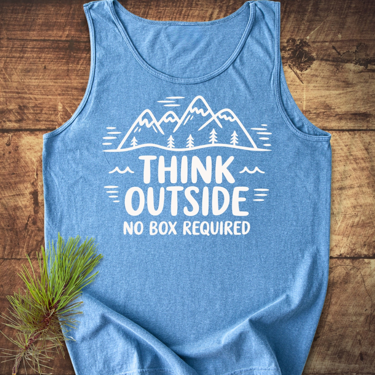Think Outside Tank Top featuring a mountain and tree design with the slogan “THINK OUTSIDE, NO BOX REQUIRED” in white, laid on wood with a pine branch accentuating its eco-friendly theme.