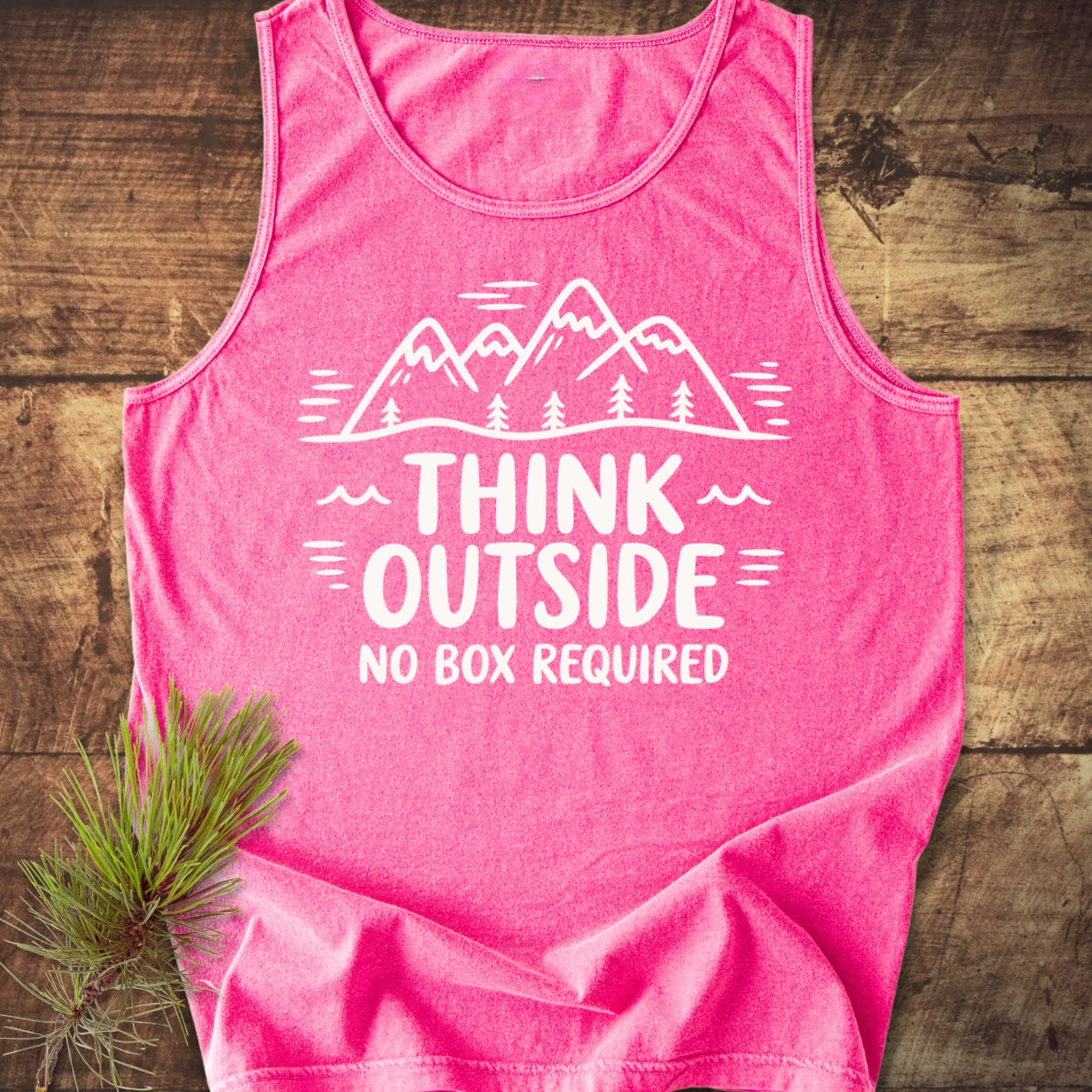 Think Outside Tank Top on wooden surface, featuring a white mountain-and-tree graphic with the slogan THINK OUTSIDE NO BOX REQUIRED, accompanied by a small pine branch beside the shirt.
