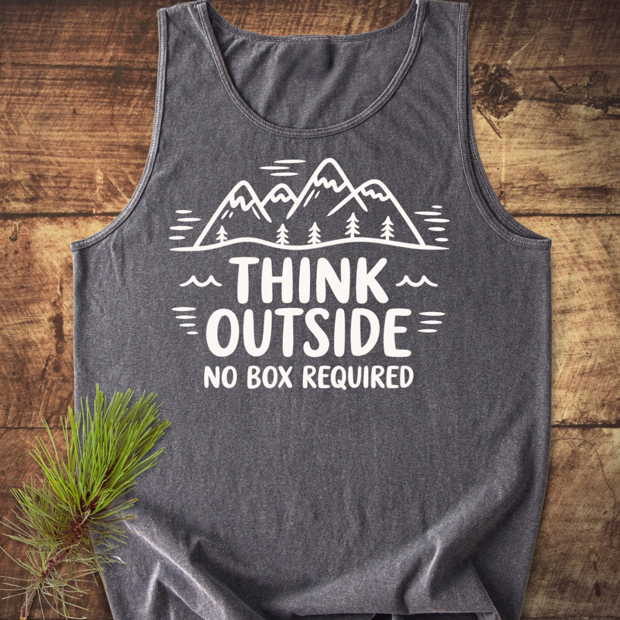 Think Outside Tank Top on rustic wood, featuring a white mountain-and-tree illustration with the slogan THINK OUTSIDE NO BOX REQUIRED. in white, highlighting its relaxed fit and sustainable, ethical production.