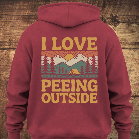 I Love Peeing Outside Hoodie BACKPRINT features a retro camping graphic with mountains, trees, and a tent. The mustard-yellow text adds humor, enhancing the garment-dyed, relaxed unisex fit for outdoor enthusiasts.