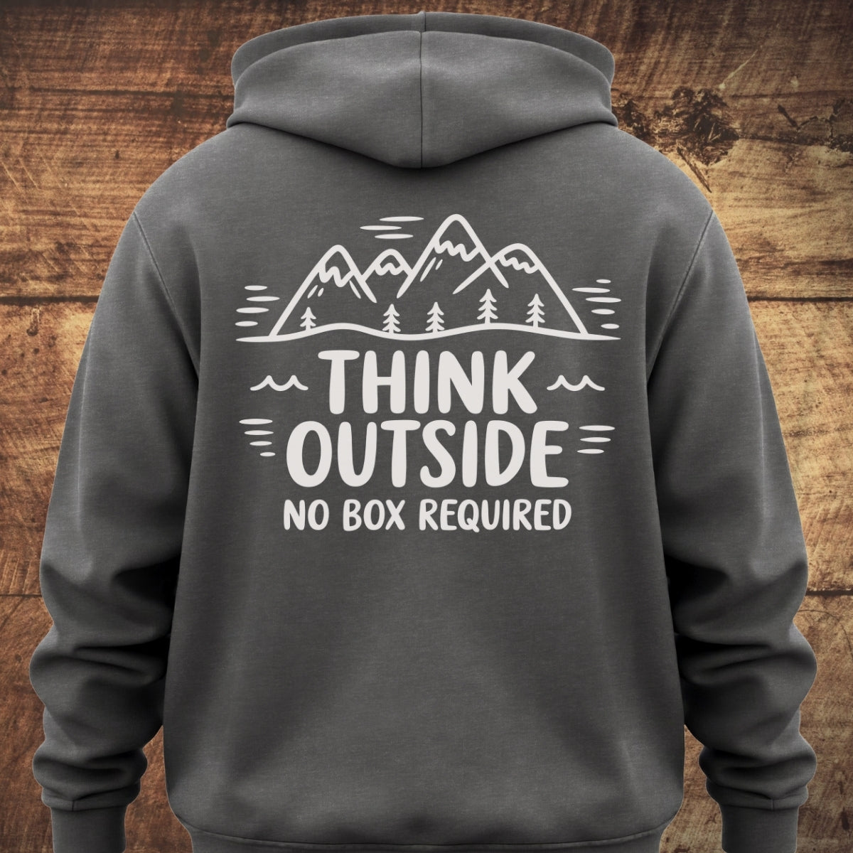 Back view of the Think Outside Hoodie BACKPRINT, showcasing a white outdoors-themed graphic with stylized mountains, pine trees, and the slogan THINK OUTSIDE NO BOX REQUIRED on a dark-gray garment-dyed unisex hoodie.