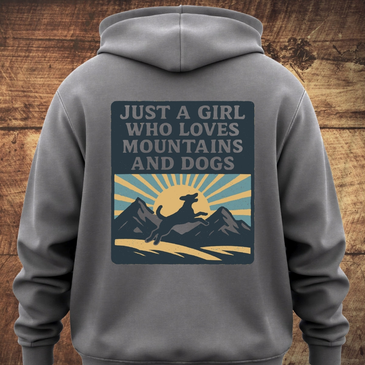 Back view of Just A Girl Who Loves Dogs Hoodie BACKPRINT, featuring a graphic of a dog leaping in front of mountains and sun. Displayed on a wooden background, showcasing its cozy, relaxed fit.