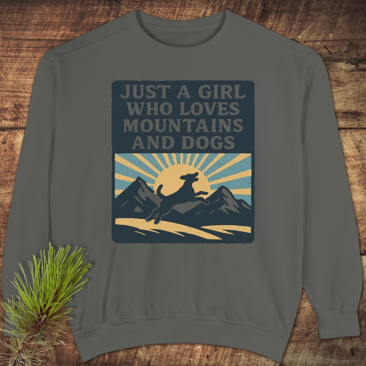 Just A Girl Who Loves Mountains And Dogs Sweatshirt on a wooden surface, featuring a retro mountain and dog graphic. The sweatshirt offers luxurious comfort with its relaxed fit and garment-dyed fleece.