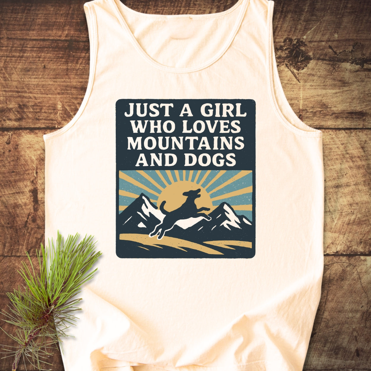 Just A Girl Who Loves Dogs Tank Top featuring a retro graphic of mountains and a dog silhouette, laid on wood with a pine sprig, embodies comfort and eco-friendly design in US cotton.