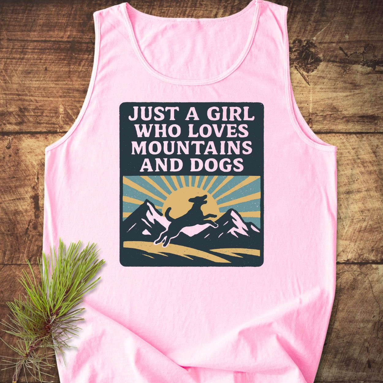 Just A Girl Who Loves Dogs Tank Top features a vintage graphic of a leaping dog, mountains, and sun, embodying a love for nature. Laid on wood with a pine branch accent.