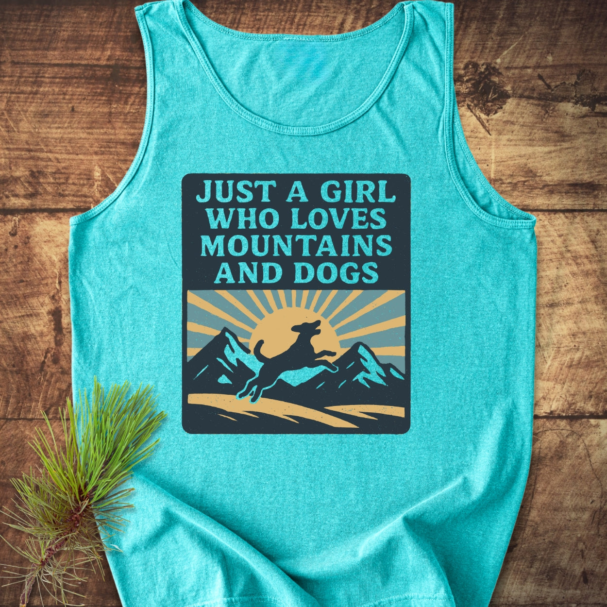 Just A Girl Who Loves Dogs Tank Top features a graphic with a dog, mountains, and sun on a teal fabric, highlighting its relaxed fit and sustainable material on a wooden surface.