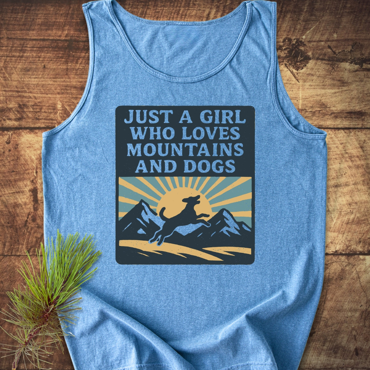 Just A Girl Who Loves Dogs Tank Top featuring a sunrise and mountain graphic with a leaping dog, laid on wood beside a pine branch, highlighting its relaxed fit and eco-friendly design.