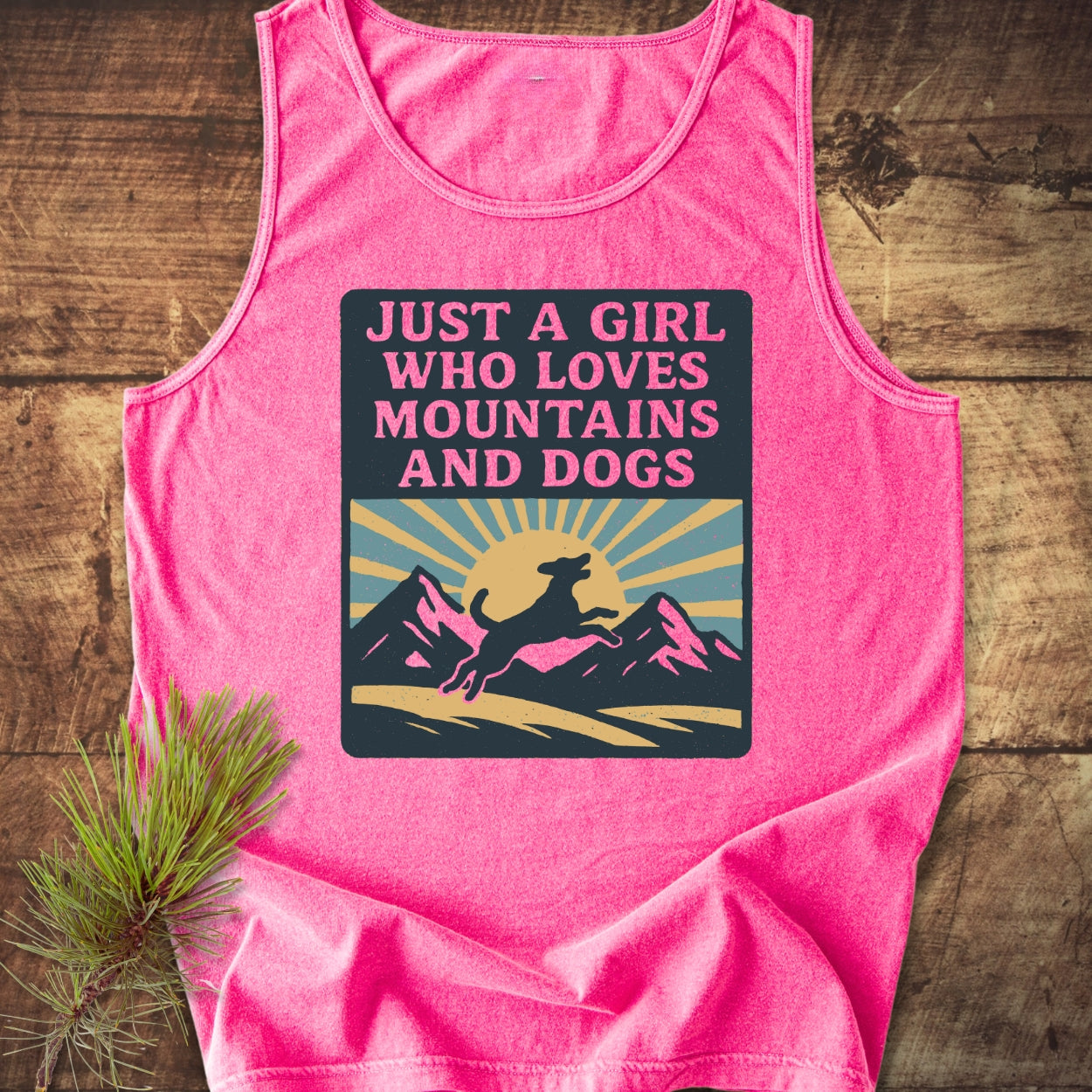 Just A Girl Who Loves Dogs Tank Top features a sunrise graphic with mountains and a dog silhouette, laid flat on wood, capturing its relaxed fit and commitment to eco-friendly style.