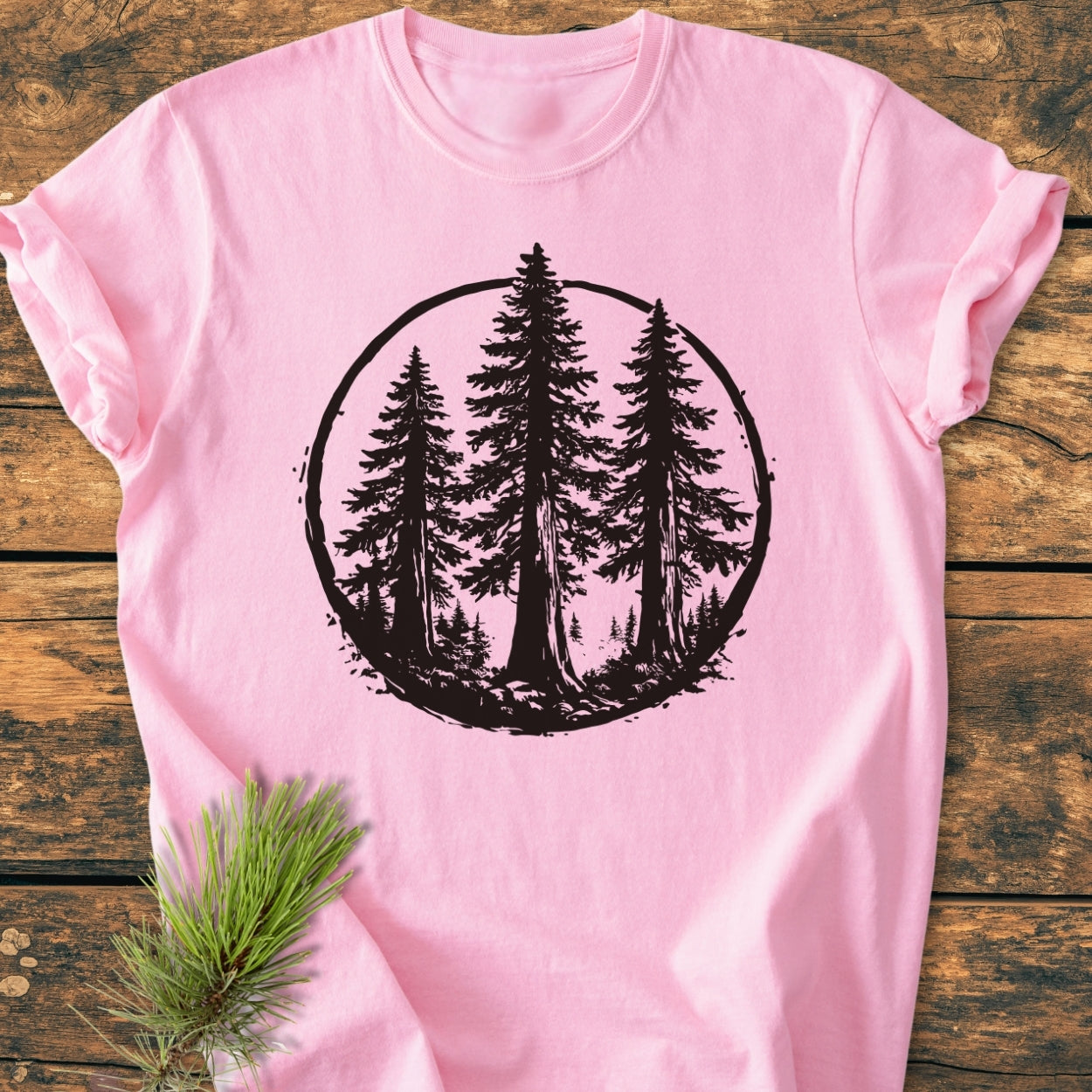 Redwood Trees - Vintage Wash Tee features a black circular graphic of evergreens on a light-pink T-shirt, laid on rustic wood. A real pine branch accentuates the shirt's nature-inspired theme.