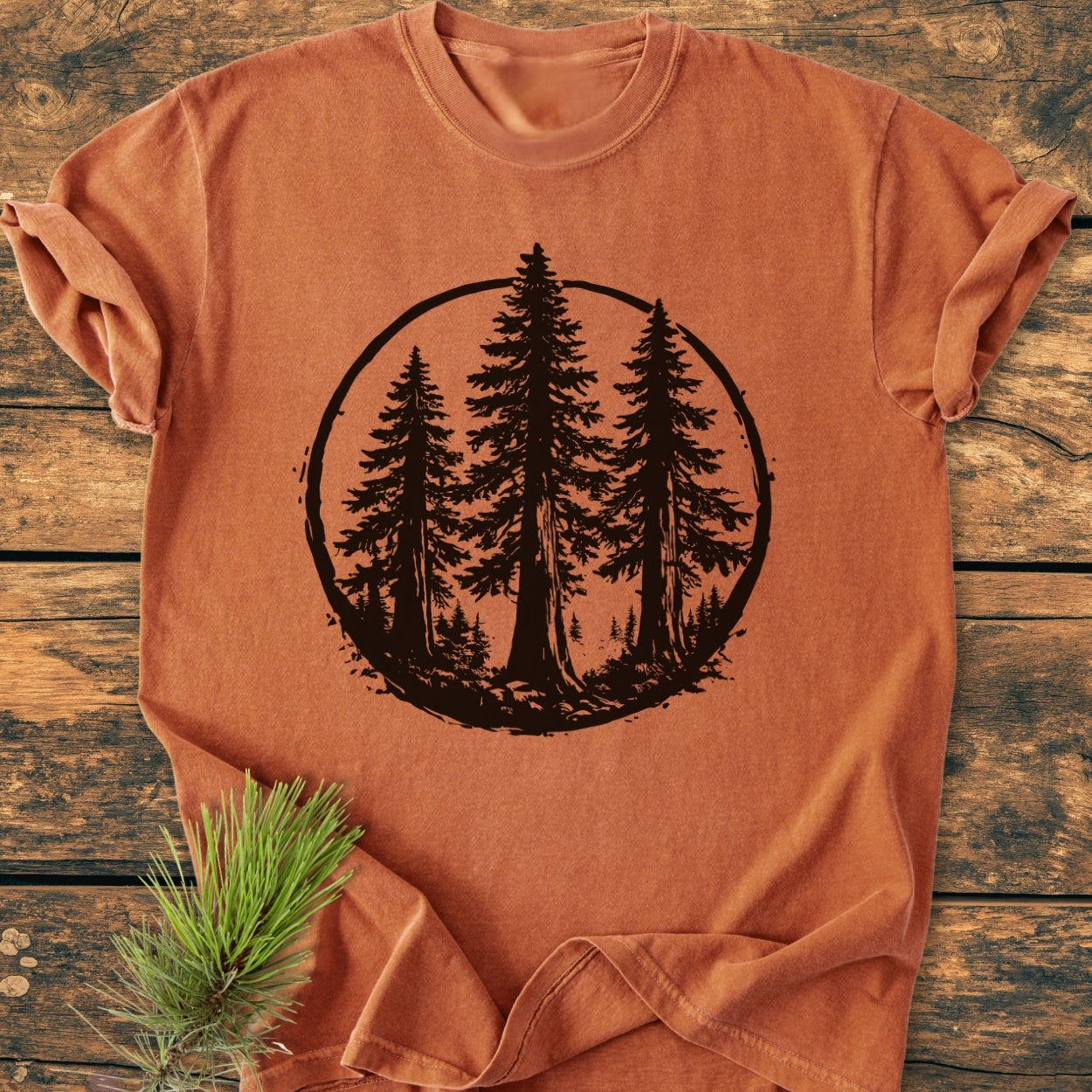 Redwood Trees - Vintage Wash Tee, a burnt-orange shirt, features a black circular graphic of tall pine trees. It’s styled on a wood surface with pine sprigs, highlighting its nature-inspired design.