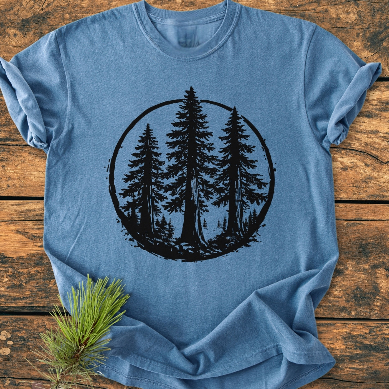 Redwood Trees - Vintage Wash Tee features a black silhouette of three pine trees within a circular frame on a soft-blue, short-sleeve T-shirt, styled with a pine bough on rustic wood.