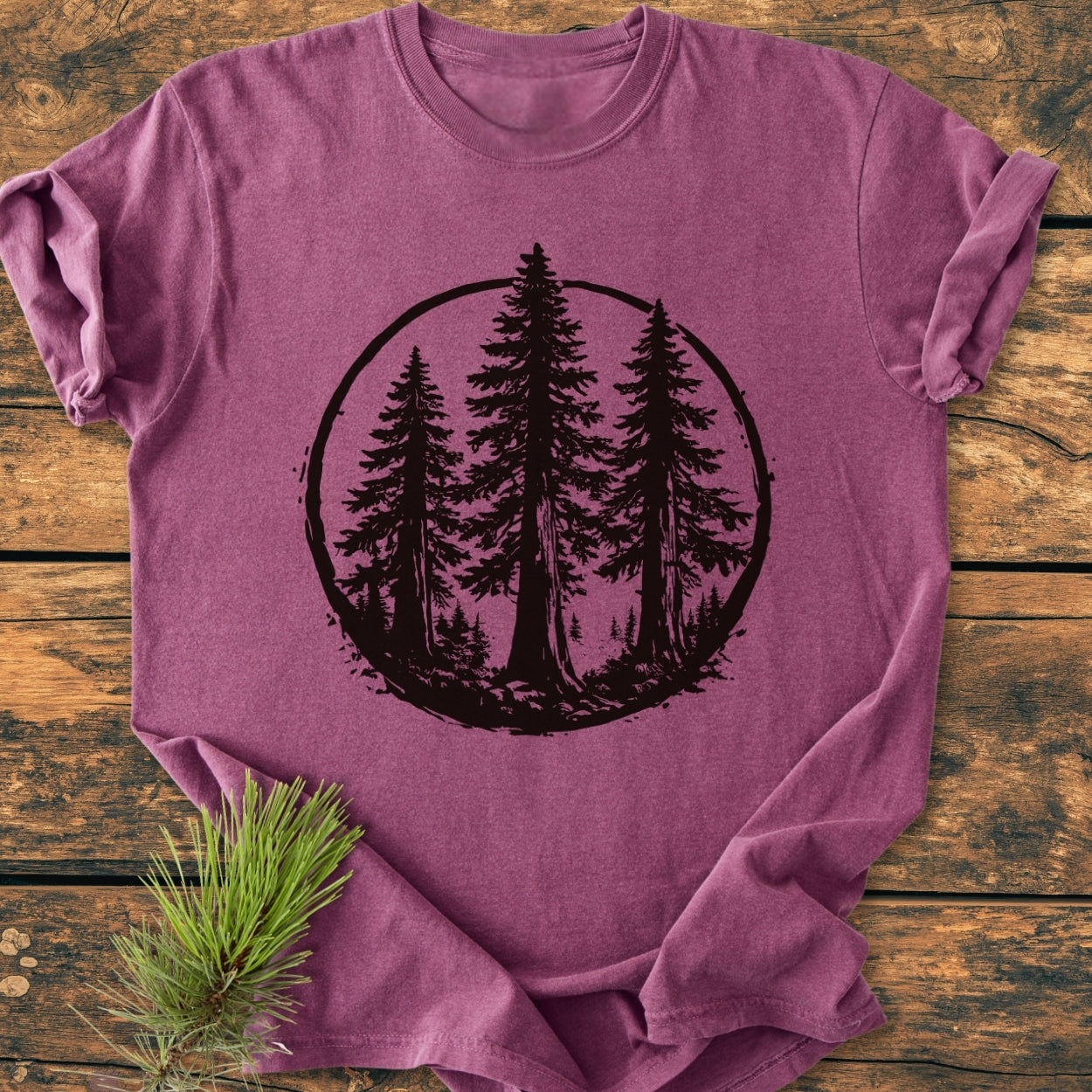 Redwood Trees - Vintage Wash Tee featuring a black silhouette of pine trees on a soft, maroon-purple t-shirt, laid flat with a pine sprig beside it, highlighting its cozy, relaxed fit.