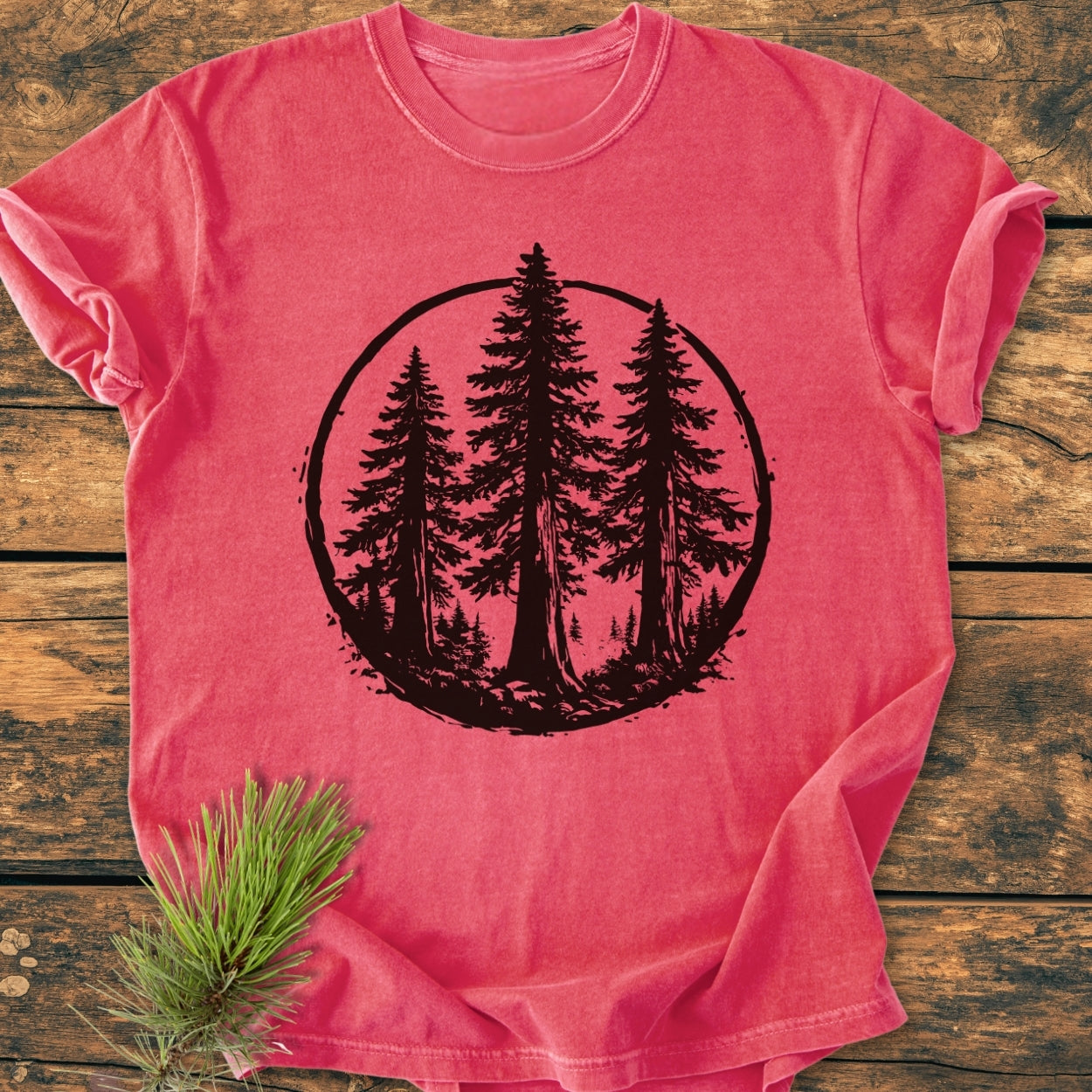 The Redwood Trees - Vintage Wash Tee features a black pine forest silhouette on a reddish-pink backdrop, laid flat on a wooden surface, with a real pine sprig accentuating its natural theme.
