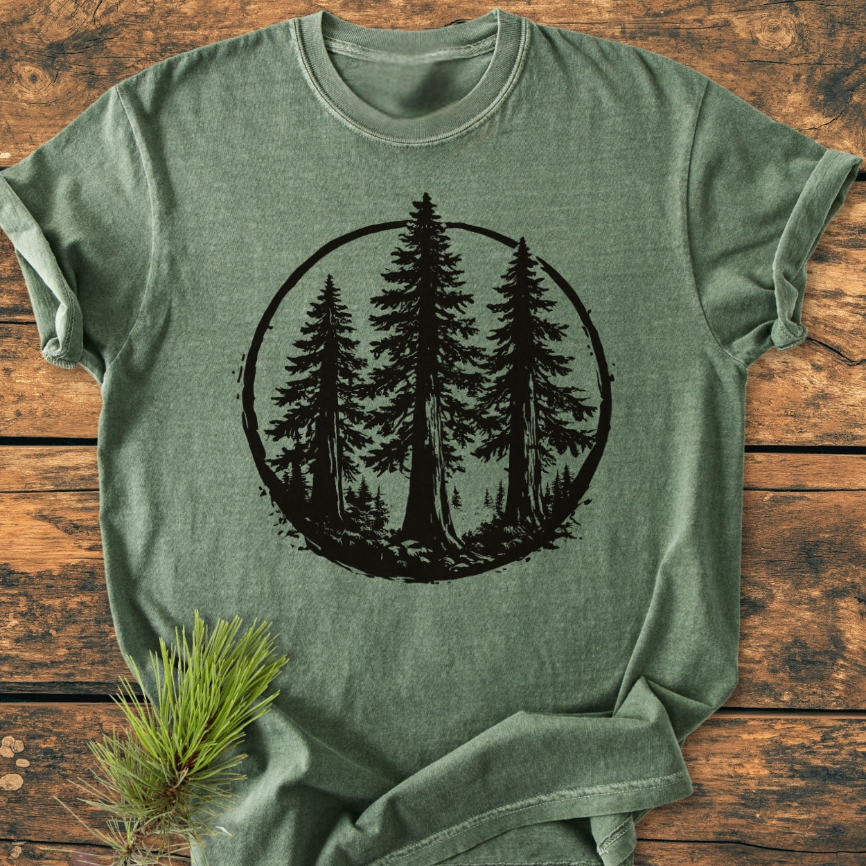 Redwood Trees - Vintage Wash Tee in heather-green, featuring a black circular graphic with three pine trees, laid flat on rustic wood with a pine sprig accent. Relaxed fit, soft-washed, durable.
