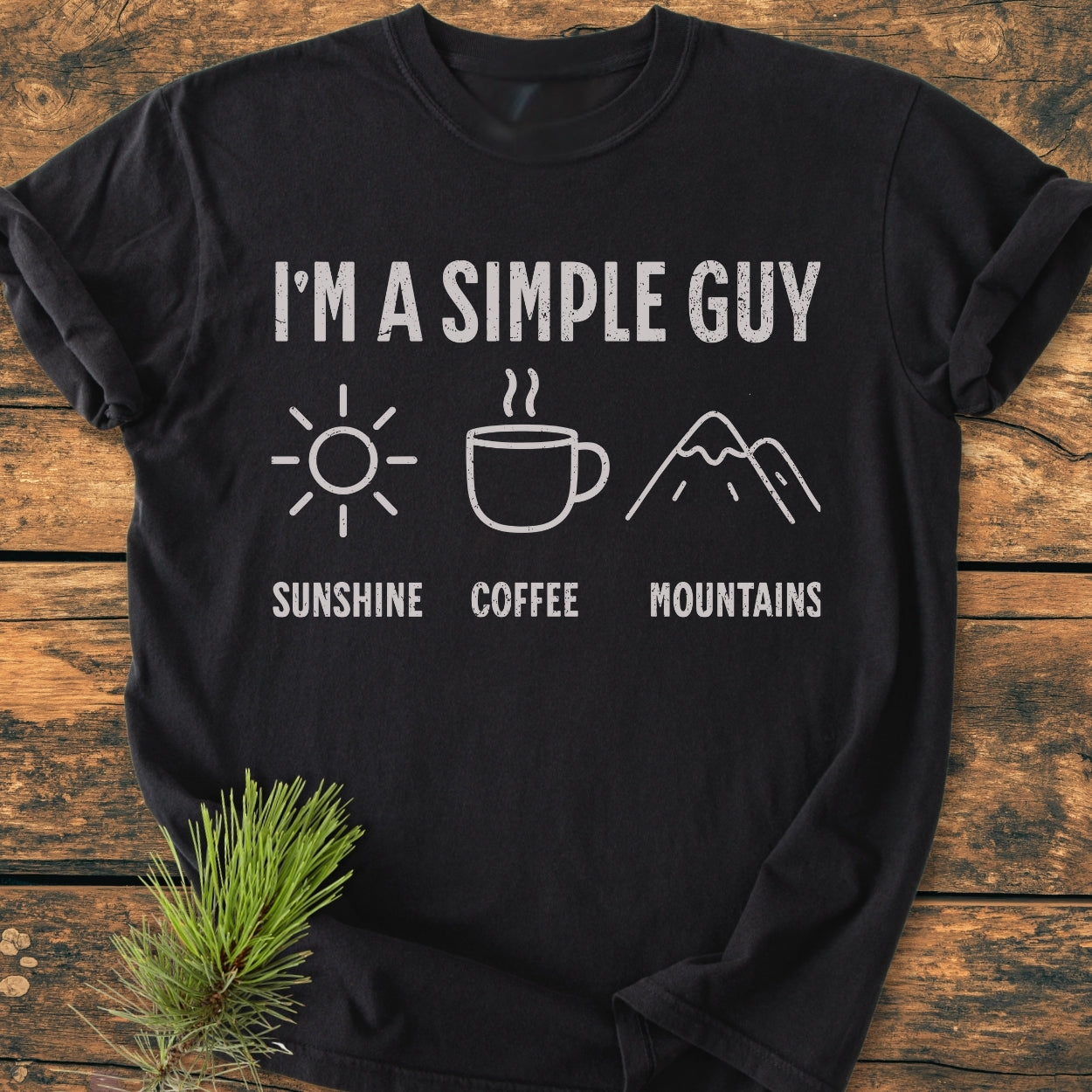 I'm A Simple Guy - Sunshine Coffee Mountains - Vintage Wash Tee features white text and icons on a black T-shirt, highlighting a sun, coffee mug, and mountains, laid on a wooden surface.