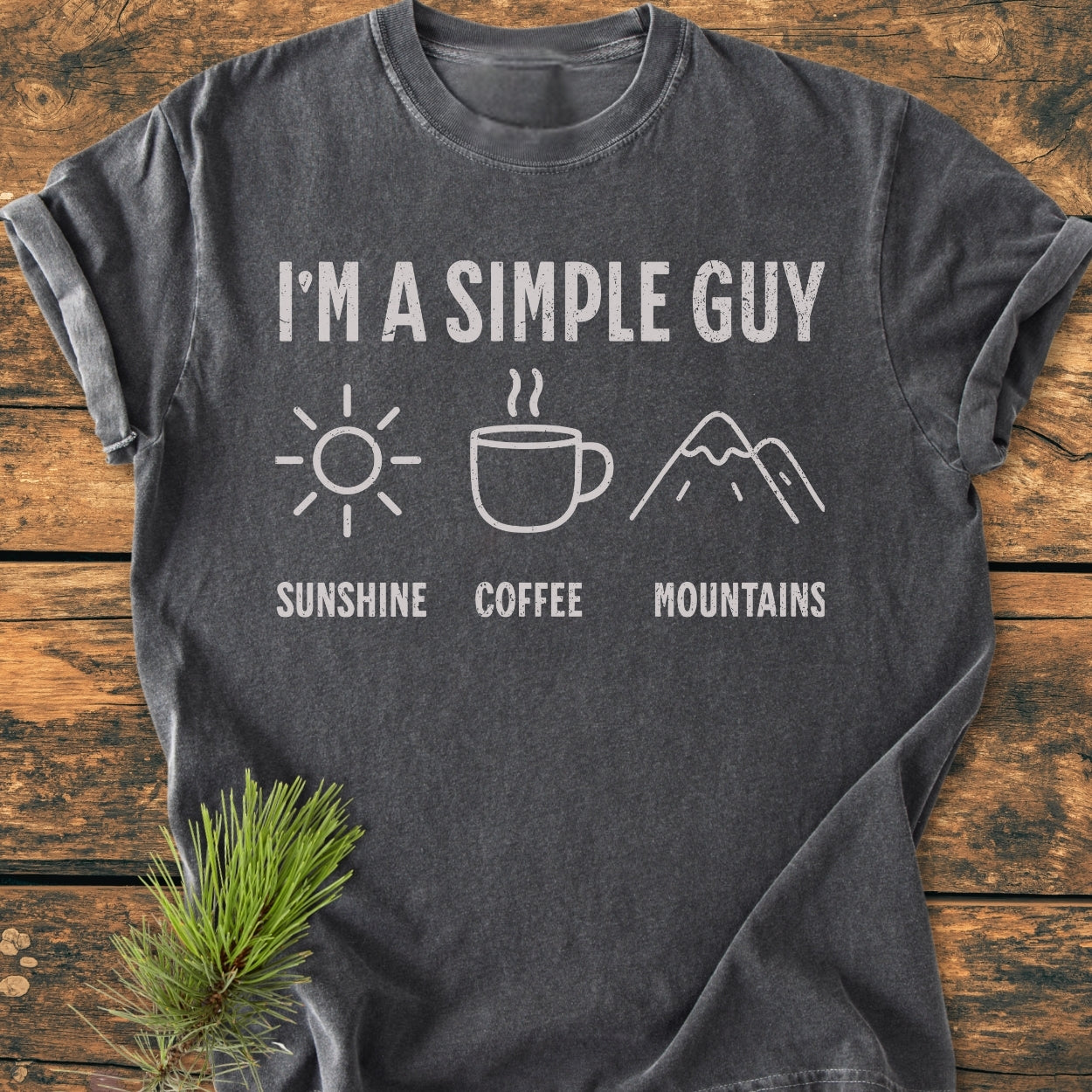 I'm A Simple Guy - Sunshine Coffee Mountains - Vintage Wash Tee displayed on a wooden surface, featuring white text and icons of a sun, coffee mug, and mountains, next to a pine sprig.