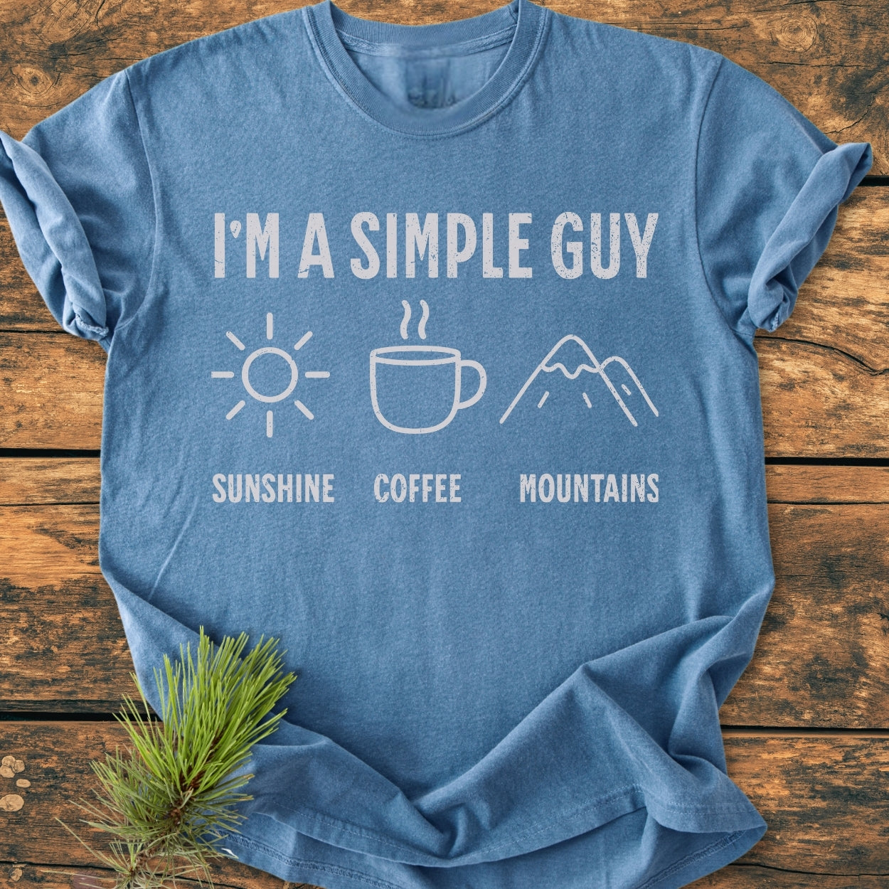 I'm A Simple Guy - Sunshine Coffee Mountains - Vintage Wash Tee in heather-blue, featuring white lettering and icons of a sun, coffee mug, and mountains, laid on wooden surface with pine sprig.