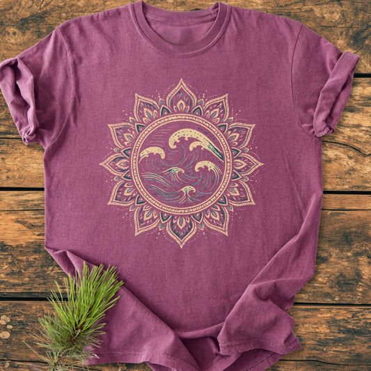 Ocean Mandala - Vintage Wash Tee laid on wood, featuring a large circular graphic of stylized waves within a sun-like border. A pine sprig rests on the heathered purple, soft-washed fabric.