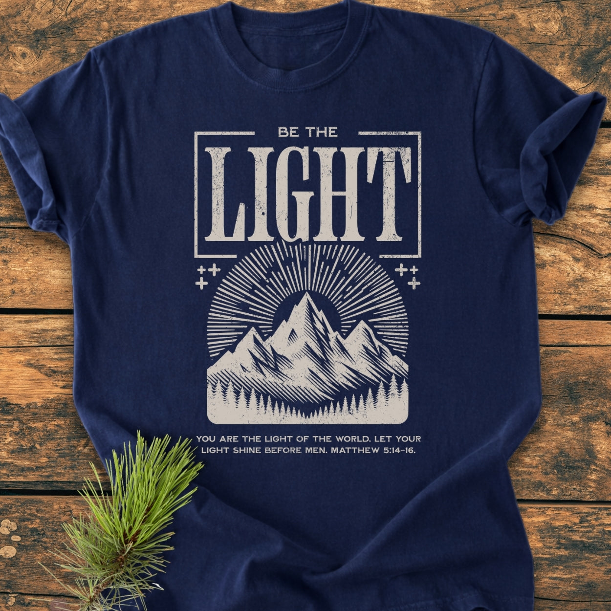 Be The Light - Vintage Wash Tee featuring a mountain range graphic with sunlight rays and pine trees, accompanied by the Bible verse Matthew 5:14–16, displayed on a wooden surface.