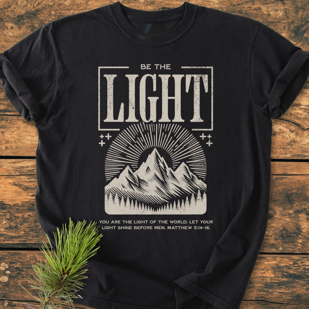 Be The Light - Vintage Wash Tee with off-white print featuring a bold headline, mountain graphic, and Bible verse reference, laid flat on a wooden surface with a pine sprig beside it.