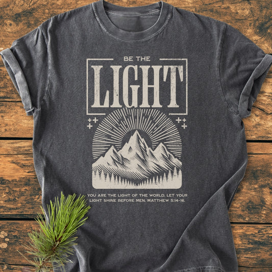 Be The Light - Vintage Wash Tee featuring a mountain and sun-ray print with Bible verse, laid flat on wood. Soft, relaxed fit, durable double-needle stitching, perfect for daily wear.