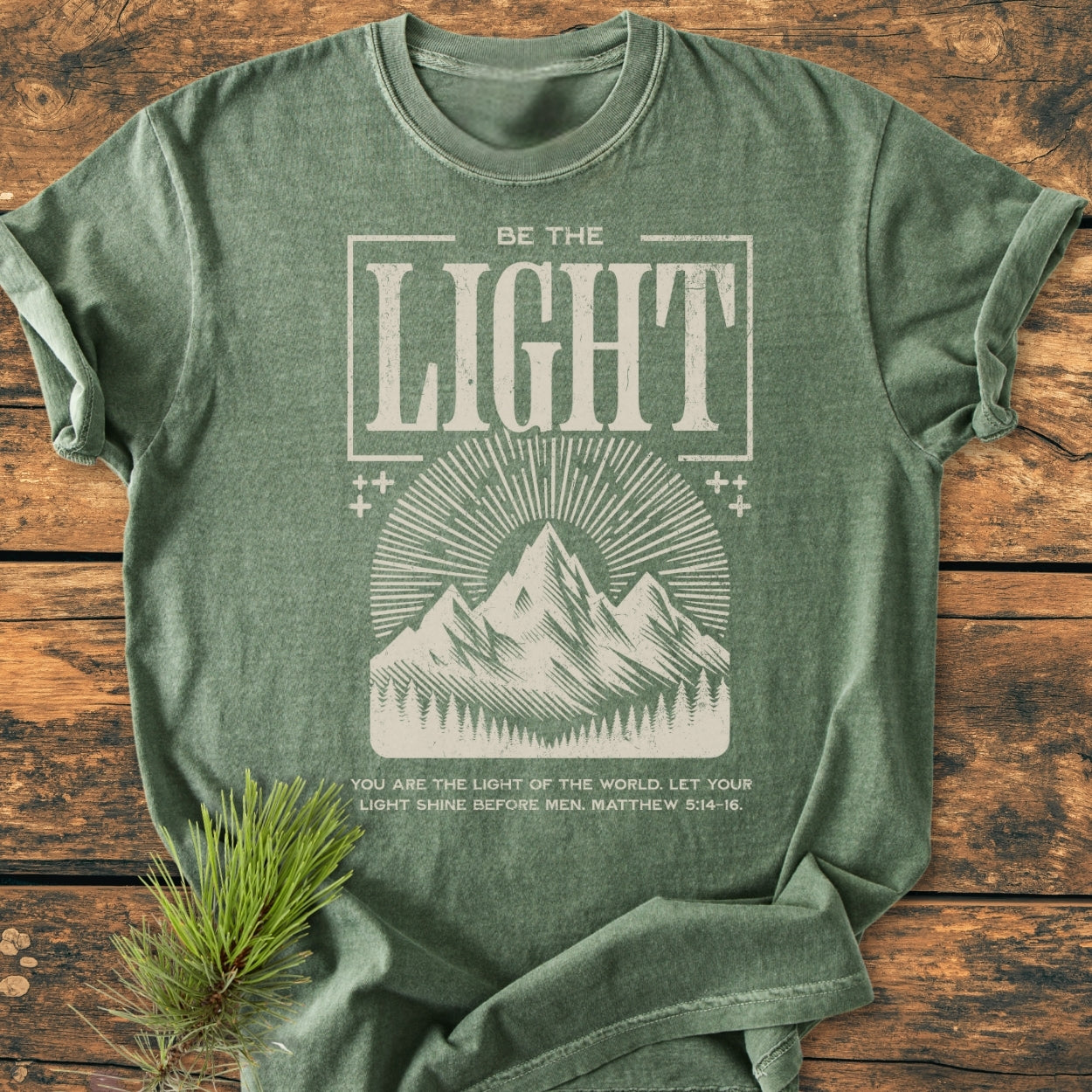 Be The Light - Vintage Wash Tee in forest-green features an off-white print of mountains, sun-rays, and pine trees with a Bible verse. Displayed flat with a pine sprig accent.