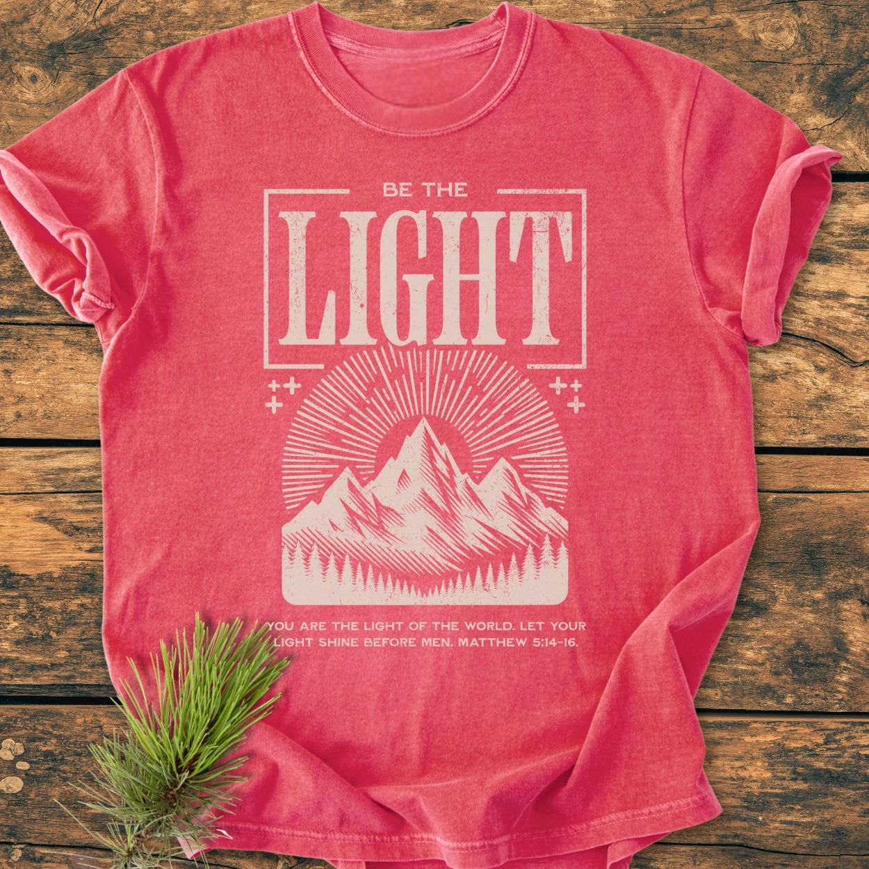 Be The Light - Vintage Wash Tee: Pink-red T-shirt with BE THE LIGHT and a sunburst design over mountains. Features Matthew 5:14–16 verse, laid on wood with a pine sprig beside.
